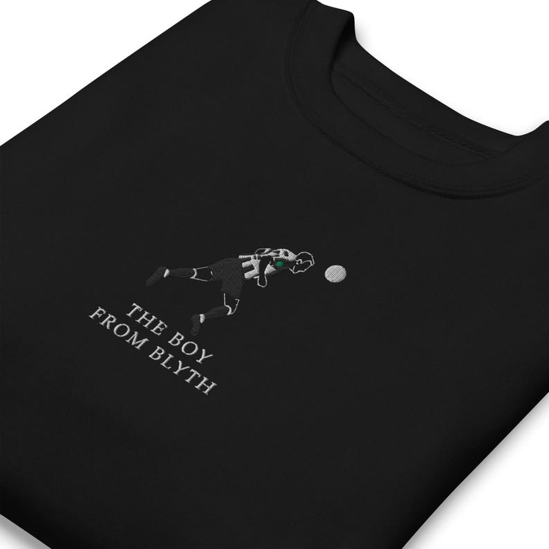 K&ouml;nnte beinhalten: Schwarzes Sweatshirt mit gesticktem Fu&szlig;ballspieler-Design. Der Text "THE BOY FROM BLYTH" ist in Wei&szlig; gestickt. Der Spieler tr&auml;gt ein schwarz-wei&szlig;es Trikot mit gr&uuml;nen Akzenten beim Schie&szlig;en.