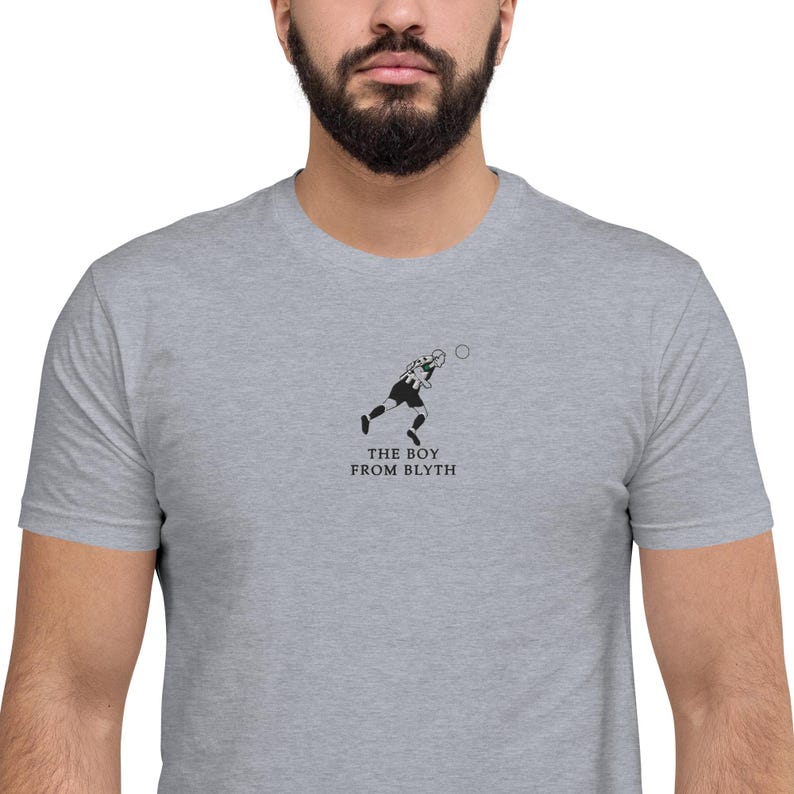 K&ouml;nnte beinhalten: Hellgraues T-Shirt mit einer minimalistischen schwarz-wei&szlig;en Illustration einer Person beim Sport und dem Text "THE BOY FROM BLYTH" darunter.