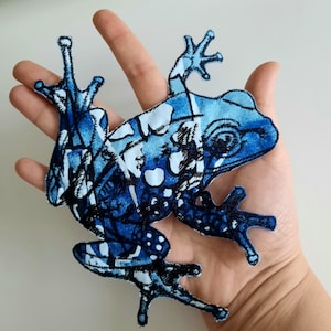 May include: A fabric patch shaped like a frog, featuring a vibrant blue and white watercolor-style pattern with dark blue and black ink-like outlines. The frog is depicted in a sprawling pose with extended limbs and detailed webbed feet.