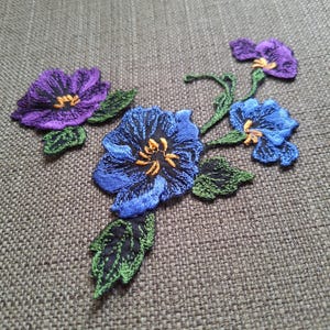 Pode incluir: Um emblema floral bordado com uma flor roxa e duas flores azuis com centros amarelos, conectadas por caules e folhas verdes. O bordado utiliza pontos detalhados para criar textura e profundidade nas pétalas e na folhagem.