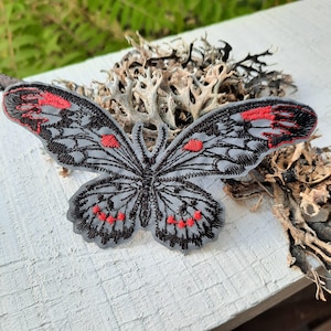 May include: An embroidered butterfly patch featuring a light grey base with intricate black thread detailing and vibrant red accents on the wing tips and lower sections. The design showcases a symmetrical pattern with detailed wing veins.
