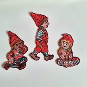 Puede incluir: Tres parches bordados con personajes festivos con gorros y trajes rojos. Una figura de pie con peto rojo y mangas a rayas, flanqueada por dos figuras sentadas con trenzas que llevan jerséis estampados y mallas a rayas.