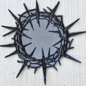 Puede incluir: Un parche circular que presenta un diseño bordado en negro de una corona de espinas. El bordado está realizado con hilo negro denso, creando una silueta afilada y puntiaguda sobre un fondo de tela gris claro con un borde troquelado.