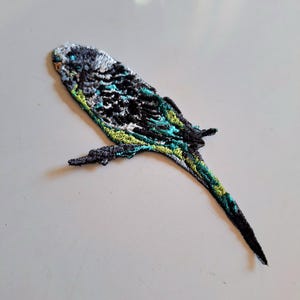 May include: An embroidered patch shaped like a budgerigar bird. The bird features a colour palette of black, white, teal, and lime green threads, with detailed stitching creating a textured appearance of feathers across its body and long tail.