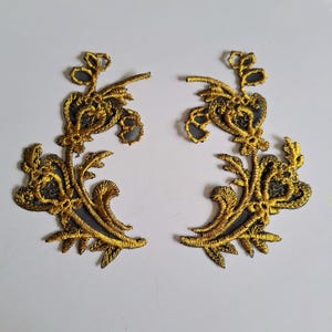 May include: Two symmetrical floral embroidery patches featuring intricate gold thread detailing on a dark grey mesh base. The design consists of curved stems, leaves, and petal shapes arranged in a mirrored pattern, showcasing dense stitching.