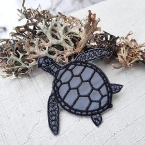 May include: A sea turtle patch featuring a smooth gray base with intricate black embroidered outlines detailing the shell and flippers. The design showcases a geometric pattern of hexagons across the carapace and detailed stitching on the limbs.