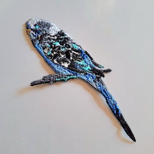 May include: An embroidered patch shaped like a budgerigar parakeet. The bird features a colour palette of black, white, and vibrant blue, with a light grey head and a long, pointed tail. The embroidery shows detailed feather patterns throughout.