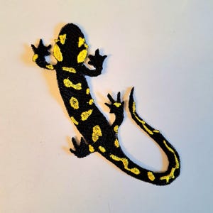 May include: An embroidered patch in the shape of a salamander. The design features a black base with bright yellow spots and markings distributed along the body, head, and tail. The creature is depicted with four legs and a long, curved tail.