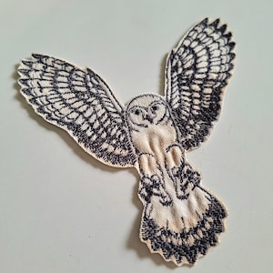 Könnte beinhalten: Ein gestickter Aufnäher, der eine fliegende Eule mit weit ausgebreiteten Flügeln zeigt. Das Design ist mit schwarzem Garn auf einem cremefarbenen Stoffgrund gearbeitet und zeigt detaillierte Federmuster an Flügeln und Schwanz sowie ein Eulen-Gesicht.