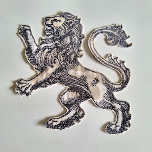 Puede incluir: Un parche bordado que presenta un león rampante en hilo azul marino oscuro sobre un fondo de tela color crema. El león está representado de perfil, de pie sobre sus patas traseras con la cola curvada hacia arriba y la boca ligeramente abierta.