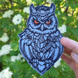 May include: A light blue embroidered patch featuring a detailed black owl design with prominent orange eyes. The owl has intricate feather patterns and a decorative band across its forehead, rendered in black thread against a light blue fabric base.