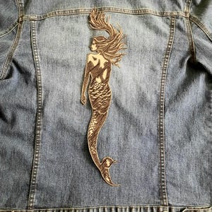May include: A blue denim jacket featuring a large, dark brown embroidered patch on the back. The patch depicts a mermaid in profile with long, flowing hair and a detailed scale pattern on the tail, rendered in a sketch-like, linear embroidery style.