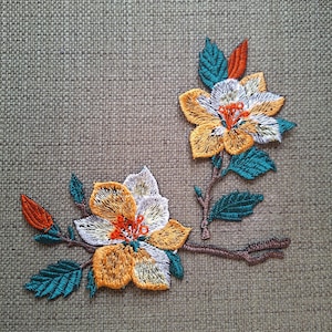 Peut inclure: Un écusson brodé représentant deux fleurs aux pétales jaune doré, blancs et bleu sarcelle, rehaussées de cœurs et de boutons orange vif. Le motif comprend des tiges brun foncé et des feuilles bleu sarcelle sur un tissu gris texturé.