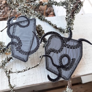 May include: Two light grey fabric patches featuring a dark black embroidered tentacle design. Each patch has a distinct black stitched border and a smooth, matte surface texture. The tentacles are depicted in a winding, curved pattern.