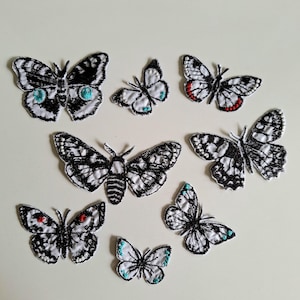 Può includere: Una collezione di otto toppe ricamate a forma di farfalle e falene di varie dimensioni. Ogni toppa presenta una base bianca con dettagli intricati in filo nero, accentuati da piccoli tocchi di rosso o turchese sulle ali.