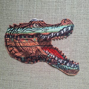 May include: An embroidered patch featuring the head of an alligator with its mouth open. The design incorporates shades of brown, orange, black, and teal, with a bright red tongue visible inside the mouth and detailed stitching throughout the scales.