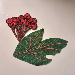 Pode incluir: Dois emblemas de tecido bordado com desenhos botânicos. Um emblema retrata um cacho de bagas vermelho-brilhantes em caules castanhos, enquanto o outro mostra uma folha verde grande de bordas serrilhadas com veios de fio dourado e rosa.
