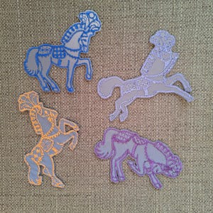 May include: Four grey fabric patches shaped like carousel horses, each featuring intricate embroidery in different colors. One horse has blue stitching, one has light purple, one has orange, and one has dark purple thread detailing.