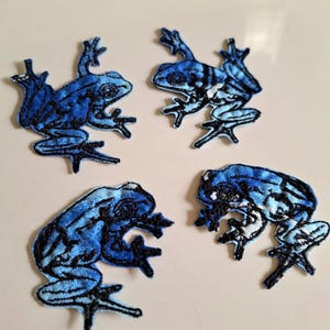 May include: Four embroidered patches shaped like frogs in various shades of blue and black. Each patch features detailed stitching to outline the frog's body, limbs, and eyes, with a gradient of light to dark blue thread creating a textured appearance.