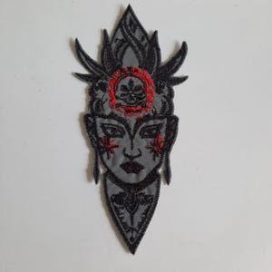 Pode incluir: Um emblema bordado apresentando um rosto estilizado com um cocar alto e pontiagudo. O design usa linha preta para os traços faciais e padrões complexos, com detalhes em linha vermelha vibrante na testa, bochechas e lábios.