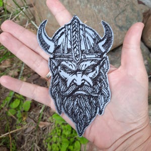 Op de afbeelding: Een lichtgrijze geborduurde patch met een gedetailleerde zwarte lijntekening van een Viking-krijger met een gehoornde helm. Het ontwerp toont een bebaard gezicht met scherpe, expressieve gelaatstrekken en ingewikkelde patronen op de helm.