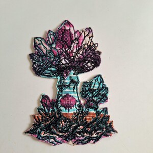 Peut inclure: Un écusson brodé représentant une forme de champignon avec un grand chapeau et une base secondaire plus petite. Le motif intègre des nuances de fils rose, violet, bleu sarcelle et noir, avec des détails complexes en forme de cristaux.