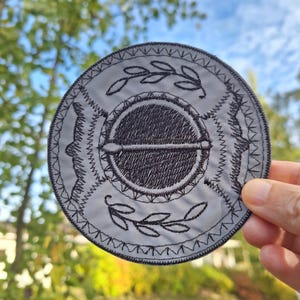 Könnte beinhalten: Ein kreisförmiger bestickter Aufnäher mit hellgrauem Hintergrund und schwarzen Faden-Details. Das Design umfasst einen dunklen Mittelkreis mit einem horizontalen Balken, umgeben von Blattmotiven und geometrischen Bordüren in Schwarz.