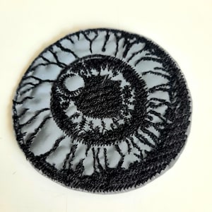 Può includere: Una toppa ricamata circolare con un design dettagliato di un occhio umano. L'iride è resa in grigio chiaro con intricate cuciture in filo nero che rappresentano la pupilla e motivi radiali che si estendono fino al bordo.
