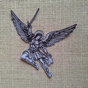 May include: An embroidered patch featuring a black line art illustration of a winged figure with a halo, depicted in a dynamic pose while holding a sword aloft. The design is rendered in dark thread against a light grey fabric background.