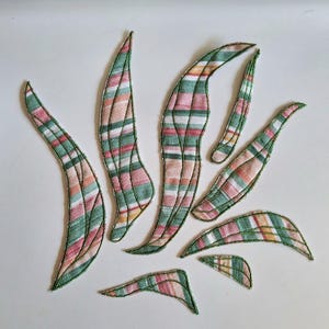 Op de afbeelding: Een verzameling van zeven bladvormige stoffen uitsparingen met een gestreept patroon in tinten groen, roze, wit en oranje. Elk stuk is afgewerkt met een decoratieve groene gestikte rand, wat een textuur geeft aan deze textielvormen.