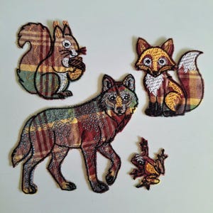 May include: Four embroidered fabric patches featuring woodland animals. The set includes a squirrel holding an acorn, a fox, a wolf, and a small frog. Each animal is crafted with a multi-coloured plaid pattern in shades of red, brown, and green.