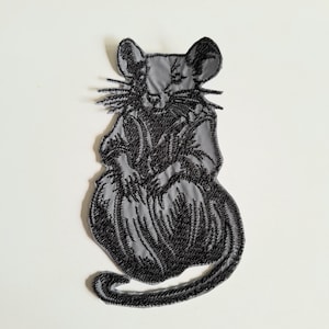 May include: A grey fabric patch featuring a detailed black embroidered design of a seated rat. The rat is depicted with prominent whiskers, pointed ears, and a long tail that curves around the base of its body, rendered in dense black thread.