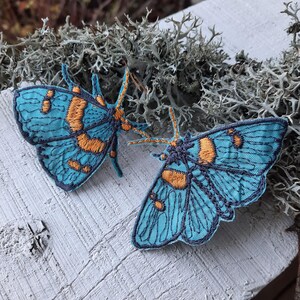 Puede incluir: Dos parches bordados de polilla con alas de color azul turquesa vibrante, contornos azul marino oscuro y detalles en naranja. Las polillas tienen antenas prominentes y patrones de hilo intrincados en sus alas, mostrando un diseño simétrico.