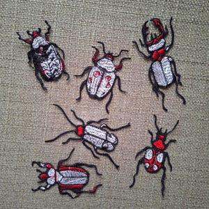 May include: A collection of five embroidered beetle patches featuring detailed stitching in shades of white, black, and red. Each insect design displays distinct anatomical features, including segmented bodies, antennae, and legs, rendered with thread.