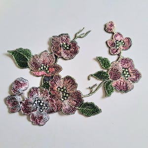 May include: An embroidered floral patch featuring multiple pink and mauve blossoms with dark green leaves. The design includes several open flowers and small buds connected by thin stems, showcasing detailed thread work and varied shading.