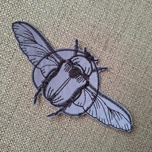 May include: A light gray fabric patch featuring a dark brown embroidered beetle design. The insect is depicted with detailed wing patterns and a segmented body, outlined with dark thread stitching against the solid light gray background.