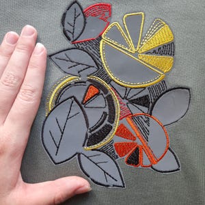 May include: A gray fabric surface featuring an embroidered design of stylized citrus fruit slices and leaves. The embroidery uses black, yellow, and orange thread to outline and fill the shapes, creating a graphic pattern on the solid gray background.