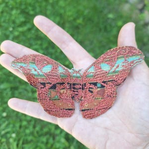Könnte beinhalten: Ein bestickter Motten-Aufnäher mit einem detaillierten Design in Rostorange, Kupfer und Türkisgrün. Die Flügel zeigen ein komplexes geometrisches Muster mit aufwendigen Stichen, die eine strukturierte Oberfläche über die gesamte Insektenform erzeugen.