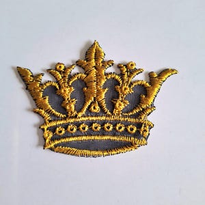 Peut inclure: Un écusson brodé en forme de couronne avec des fils dorés métallisés sur une base en maille gris foncé. Le motif présente des détails en filigrane complexes, une bande horizontale avec des accents circulaires et cinq pointes au sommet.