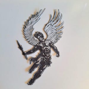 Könnte beinhalten: Ein gestickter Aufnäher mit einer geflügelten Figur im Flug, gefertigt aus schwarzem und weißem Garn. Die Figur hält einen spitzen Gegenstand in einer Hand, wobei detaillierte Stickereien die Muskulatur, fließende Gewänder und Flügel betonen.