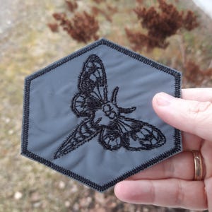 May include: A hexagonal gray patch featuring a detailed black embroidered moth design. The patch has a thick black stitched border around the entire perimeter, creating a clean edge. The moth illustration is centered on the smooth gray fabric surface.