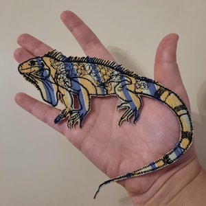 Könnte beinhalten: Ein gestickter Aufnäher in Form eines Leguans mit einem Muster aus vertikalen gelben und hellblauen Streifen. Das Echsendesign ist mit schwarzem Garn umrandet und weist komplexe schwarze Stickdetails entlang des Rückens und Schwanzes auf.