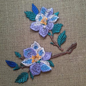 Puede incluir: Dos parches bordados de flores con pétalos de color morado claro y azul con centros de color amarillo brillante. Las flores están unidas a tallos marrones con hojas de color verde azulado oscuro. El bordado muestra una textura de hilo visible.