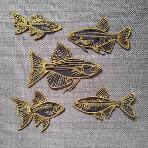 Peut inclure: Cinq écussons brodés en forme de poisson avec une base en tissu bleu marine foncé et des détails en fil doré soulignant les nageoires, les écailles et les structures squelettiques. L'ensemble comprend diverses formes de poissons.