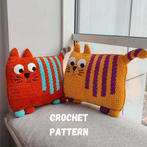 Könnte beinhalten: Zwei gehäkelte Katzenkissen. Eines ist orange mit blauen Streifen, das andere gelb mit lila Streifen. Beide haben große weiße Augen, schwarze Schnurrhaare und kleine Ohren. Der Text "CROCHET PATTERN" steht darunter.