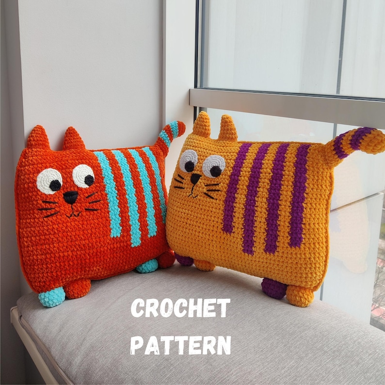 May include: Two crocheted cat cushions. One is orange with blue stripes, the other is yellow with purple stripes. Both have large white eyes, black whiskers, and small ears. The text "CROCHET PATTERN" is below the cushions.