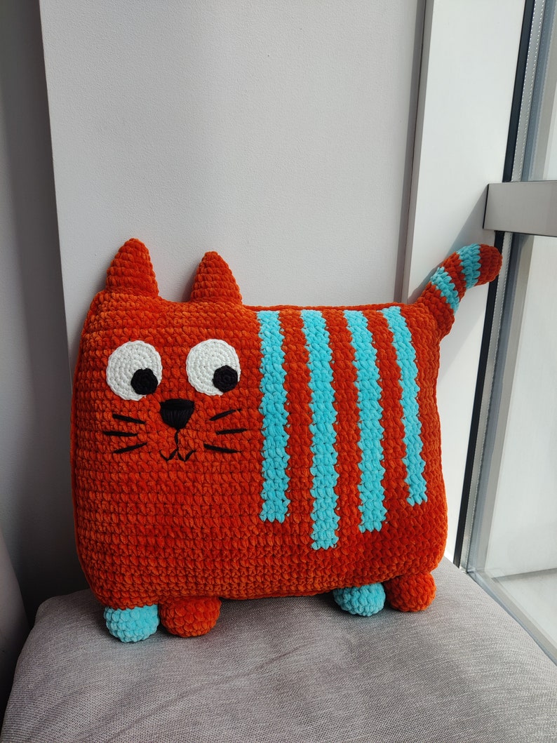 May include: A handmade orange cat-shaped cushion with blue stripes and accents. The plush cushion features a smiling face with white eyes, black pupils, and whiskers. The cat has small ears and a striped tail. The cushion is on a grey surface.
