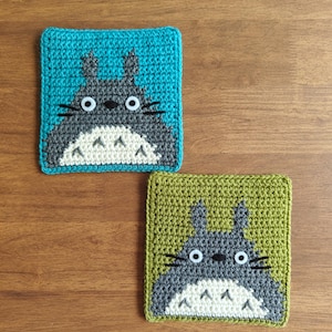 May include: Two crocheted square coasters featuring the character Totoro. One coaster has a teal background, the other a green background. Both have a gray Totoro design with white belly and eyes. The coasters are placed on a wooden surface.