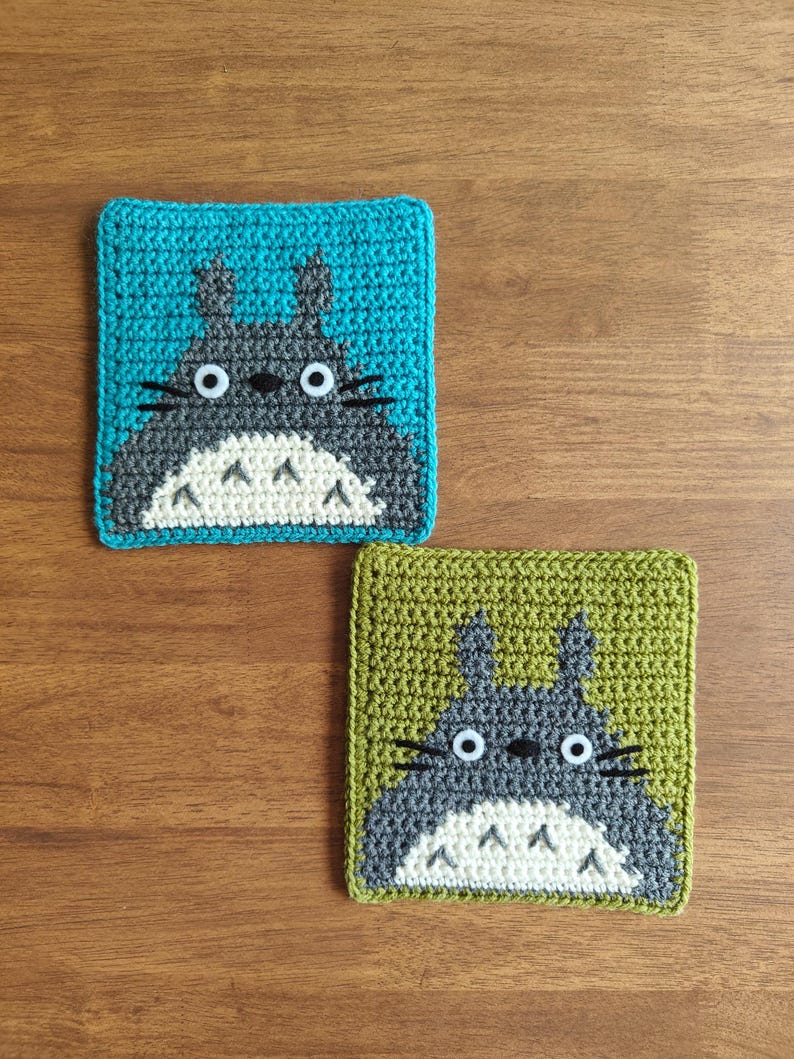 May include: Two crocheted square coasters featuring the character Totoro. One coaster has a teal background, the other a green background. Both have a gray Totoro design with white belly and eyes. The coasters are placed on a wooden surface.