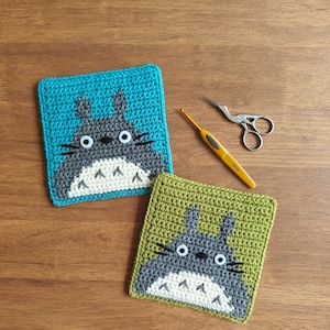 May include: Two crocheted coasters featuring a cartoon character design. One coaster is teal, the other is olive green. Both have a gray character with white accents and black whiskers. A yellow crochet hook and small scissors are also on the wooden surface.